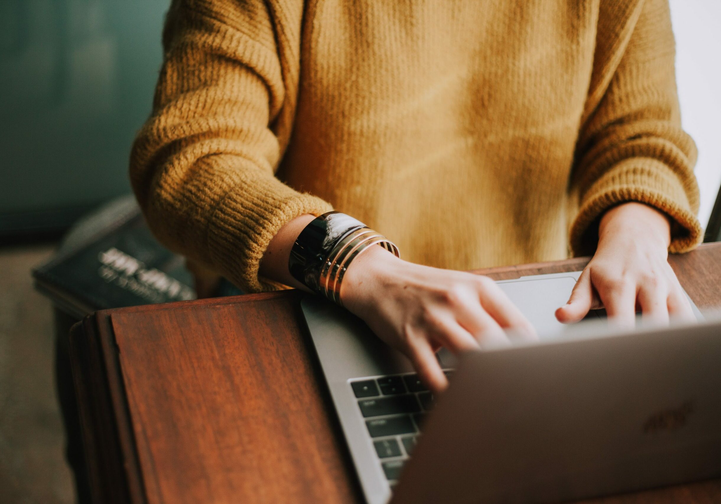 A woman in a cute camel-colored sweater and metallic bracelets, working on her laptop.