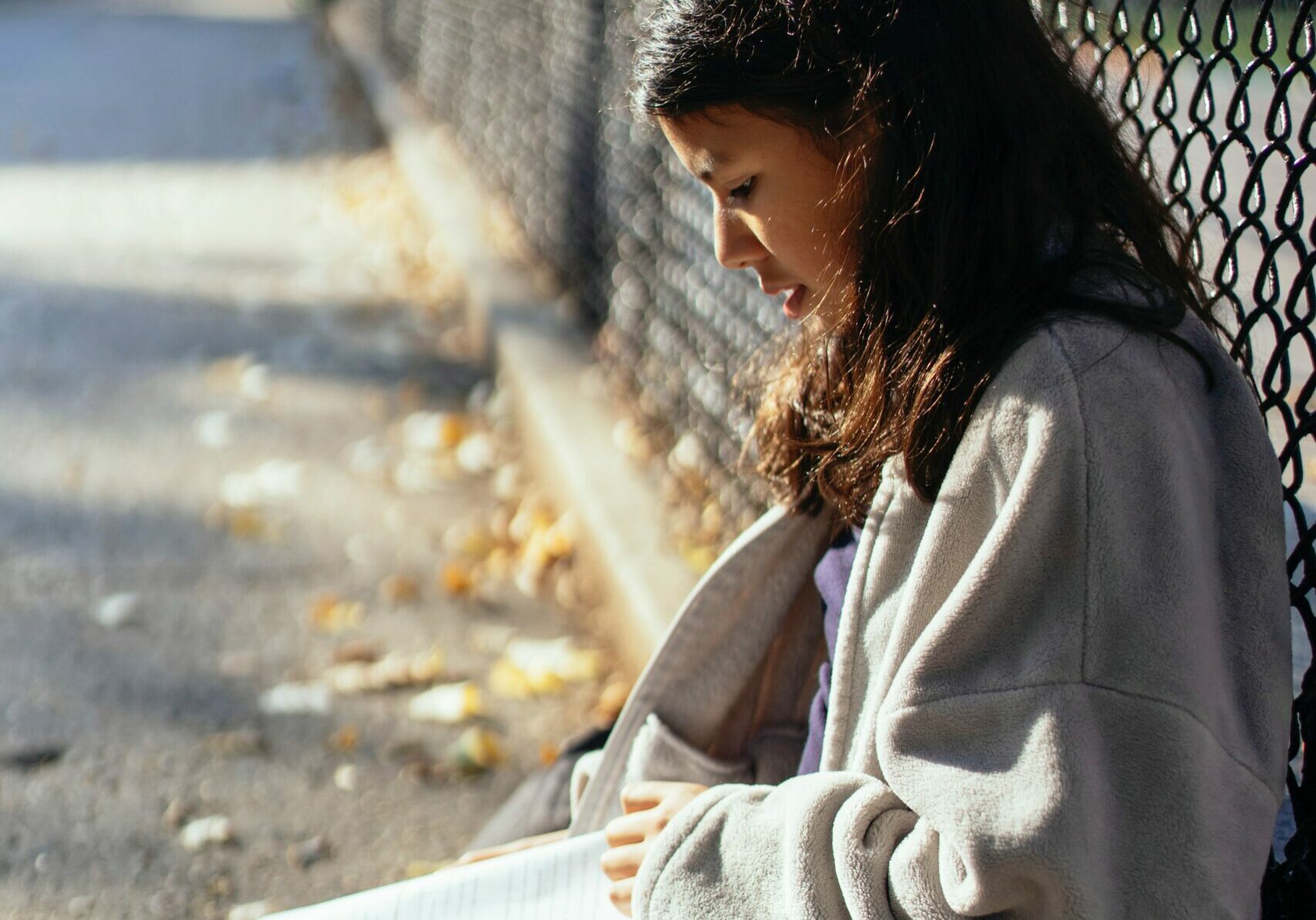 Girl sitting on the ground outside, against a fence, reading a textbook.