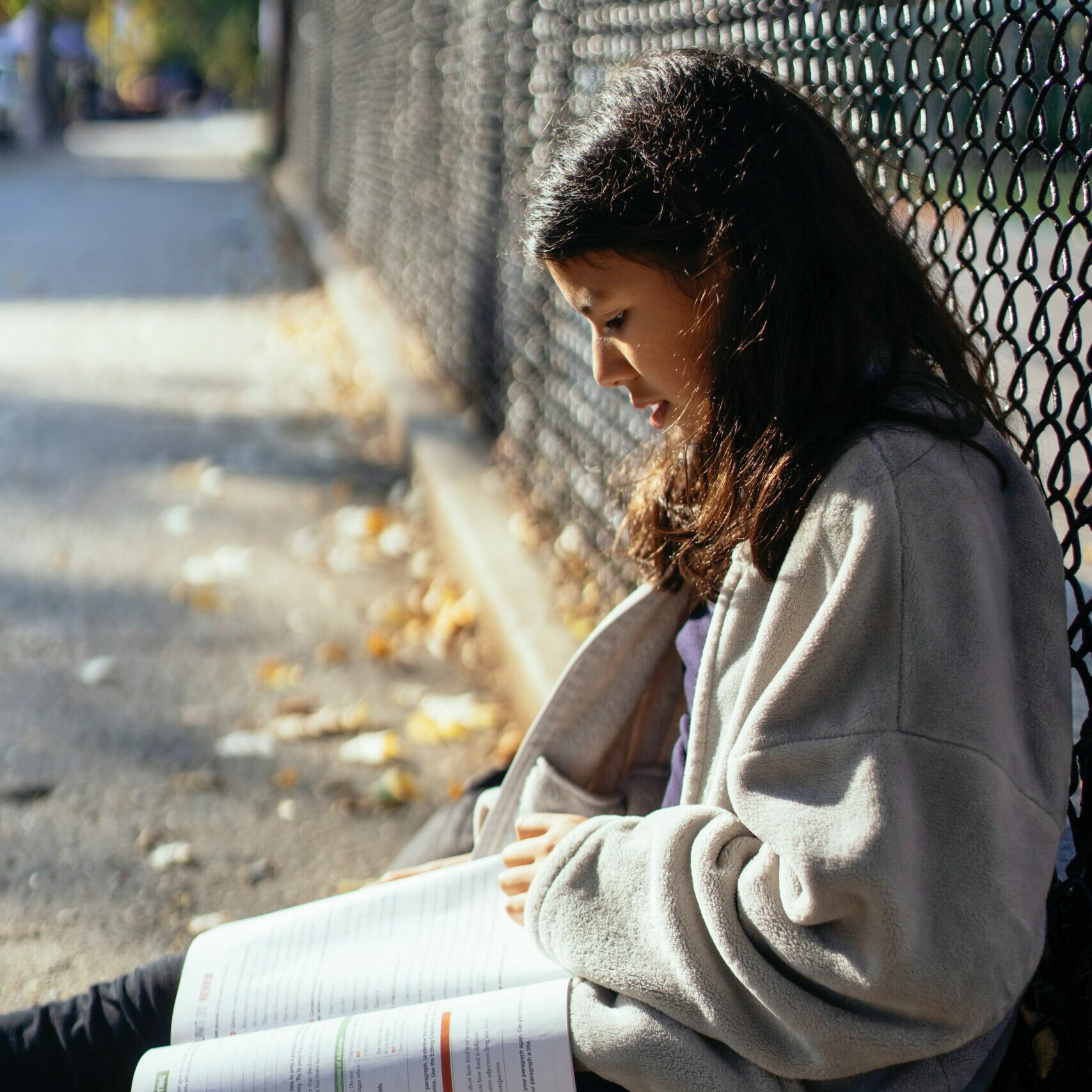 Girl sitting on the ground outside, against a fence, reading a textbook.